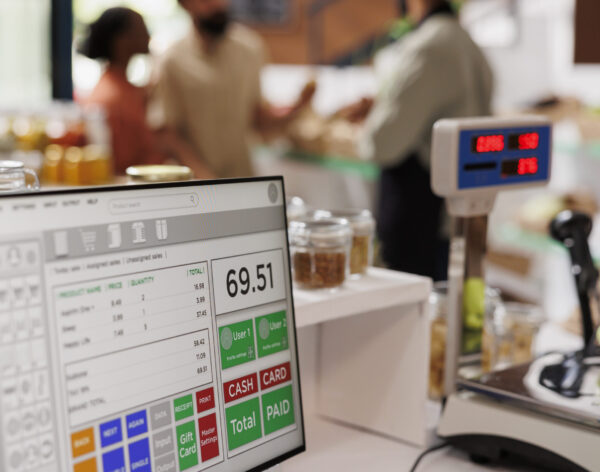 While multiethnic clients are assisted by salesman in the background, apples are placed on digital measuring scale. Photo focus on cashier's desk with weighing equipment and desktop computer.