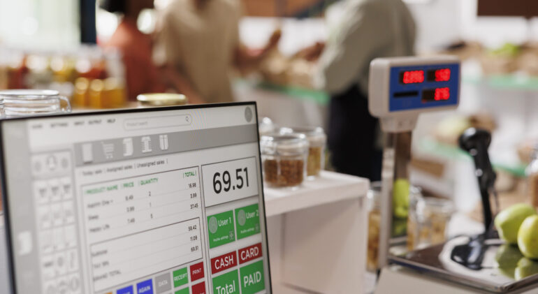 While multiethnic clients are assisted by salesman in the background, apples are placed on digital measuring scale. Photo focus on cashier's desk with weighing equipment and desktop computer.