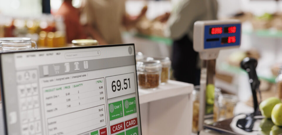 While multiethnic clients are assisted by salesman in the background, apples are placed on digital measuring scale. Photo focus on cashier's desk with weighing equipment and desktop computer.